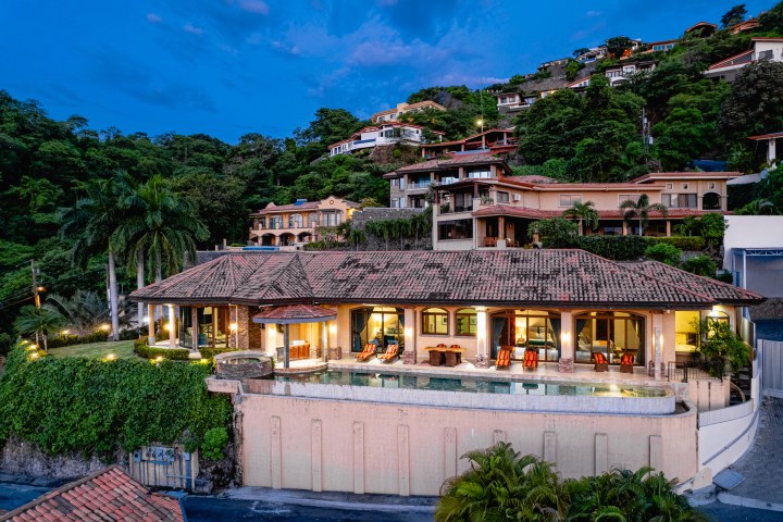 Luxurious hillside villa with pool, surrounded by lush greenery and other homes at dusk.