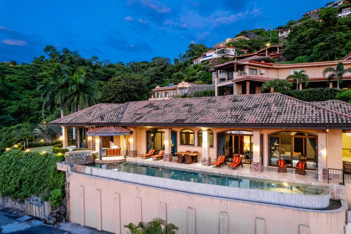 Luxurious villa with pool, surrounded by lush greenery and hilltop houses at dusk.