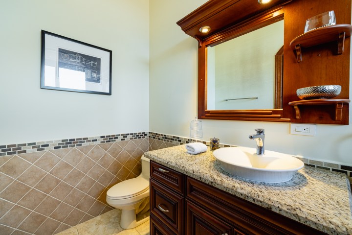 Bathroom with wood vanity, round sink, mirror, and framed art on tiled wall.