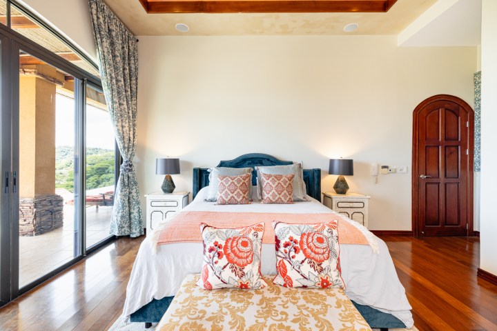 Elegant bedroom with large bed, pillows, two bedside lamps, and a window with curtain, overlooking greenery.