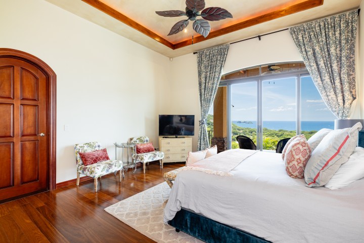 Sunny bedroom with ocean view, bed, chairs, TV, and large window.