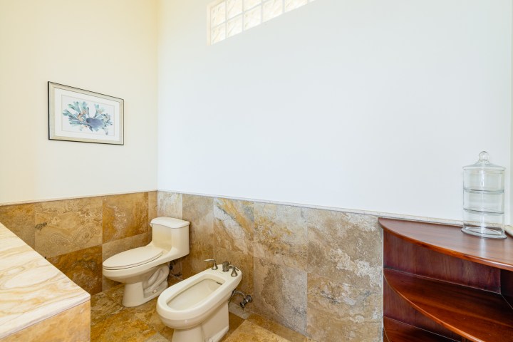 Bathroom with toilet, bidet, artwork, and glass jar on wooden shelf.