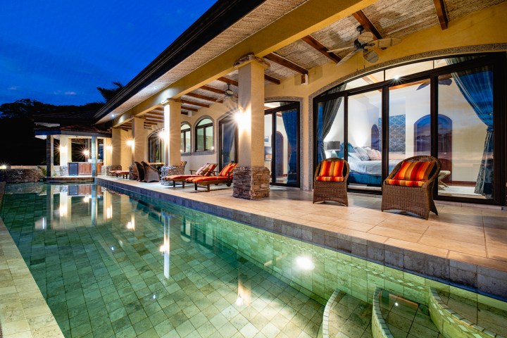 Luxury villa with pool, lounges, and lit interior at night.