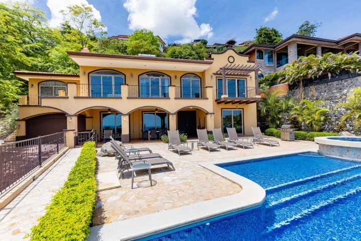 Luxurious house with large windows, outdoor pool, and patio chairs, surrounded by greenery.