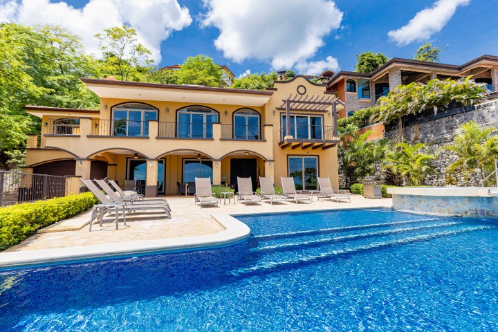 Luxury villa with a pool, surrounded by lush greenery and a bright blue sky.