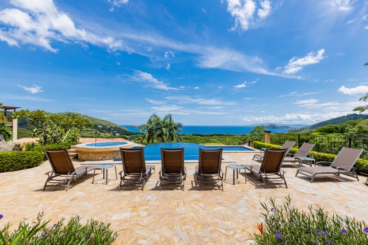 Lounge chairs by a pool with ocean view, surrounded by green hills under a blue sky.