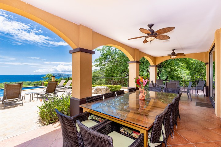 Covered patio with dining area overlooking a pool and ocean view, featuring ceiling fans and wicker furniture.