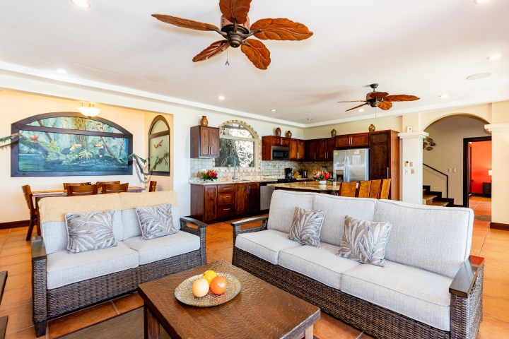 Open-plan living room with wicker sofas, kitchen, and dining table under ceiling fans and tropical decor.