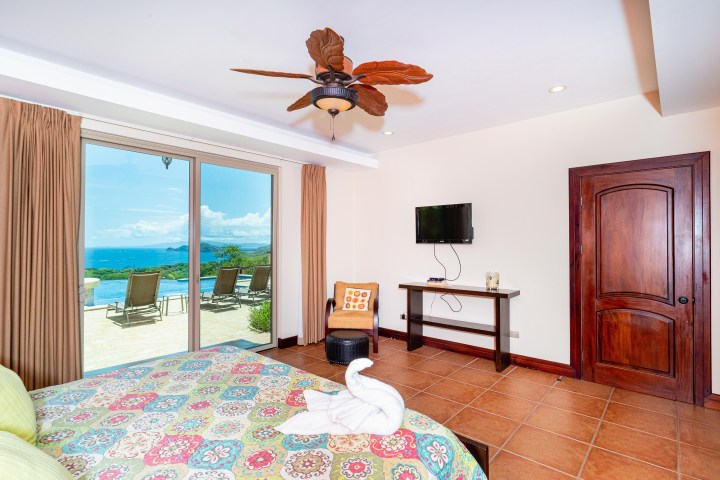 Bedroom with large window overlooking ocean, fan, TV, and colorful bedspread.