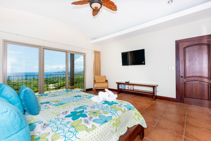 Bedroom with ocean view, floral bedding, TV, ceiling fan, and wooden door and floor.