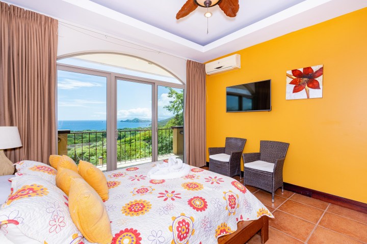 Bright room with ocean view, floral bedspread, yellow walls, TV, chairs, and balcony window.