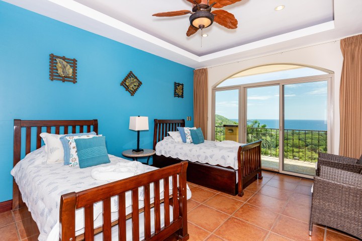 Bedroom with two beds, blue wall, ceiling fan, and ocean view through large window.