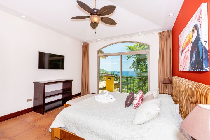 Bedroom with ocean view, red accent wall, ceiling fan, TV, and toucan artwork.