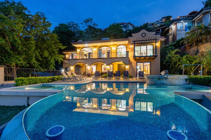 Luxury villa at dusk with illuminated pool, surrounded by trees and hillside gardens.