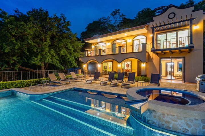 Luxury villa at night with pool, hot tub, and lounge chairs, surrounded by trees.