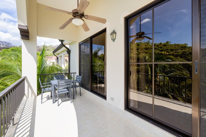 Covered balcony with table, chairs, ceiling fan, and glass doors facing green foliage.