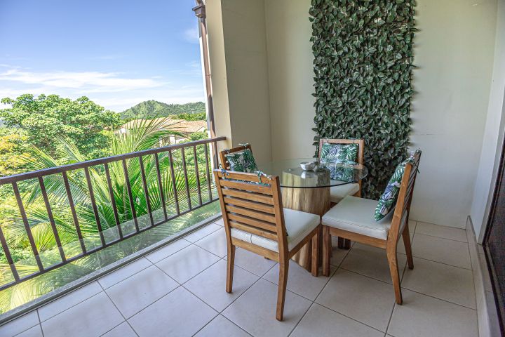 Balcony with a glass table, four chairs, and leafy view.