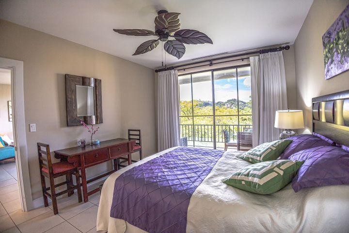 A bedroom with a bed, desk, mirror, ceiling fan, and large window with a scenic view.