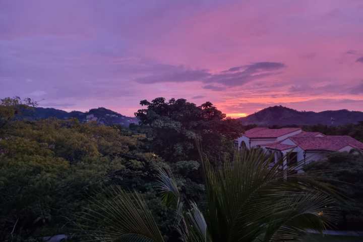 Colorful sunset over hilly landscape with trees and red-roofed house.