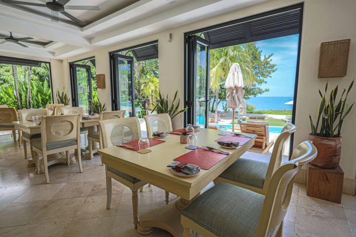 Bright restaurant interior with beach view, tables set with red mats, glass doors open to pool and sea.