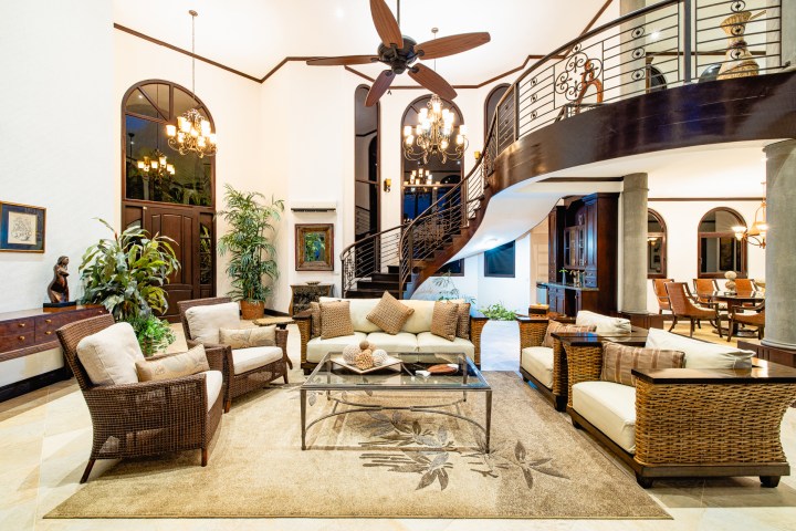 Spacious living room with wicker furniture, a glass coffee table, staircase, and chandeliers overhead.