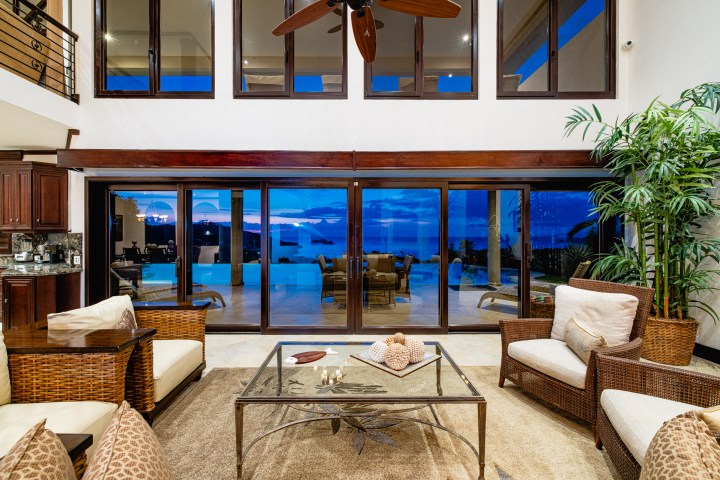 Spacious living room with wicker furniture and large windows showing a sunset view over a pool.