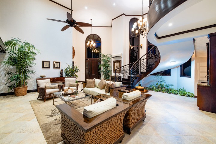 Elegant living room with spiral staircase, wicker furniture, large plants, and a chandelier.