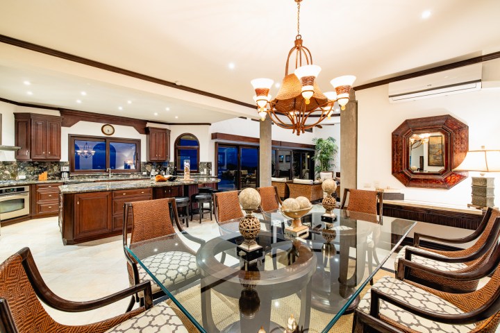 Elegant open kitchen and dining area with chandelier, glass table, and wooden cabinets.