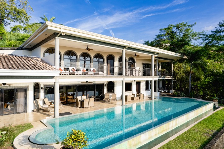 Luxury villa with arched windows, balcony, and infinity pool, surrounded by lush greenery and blue sky.