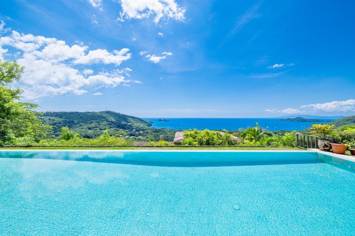 Infinity pool overlooking lush hills and blue ocean under a clear, sunny sky.