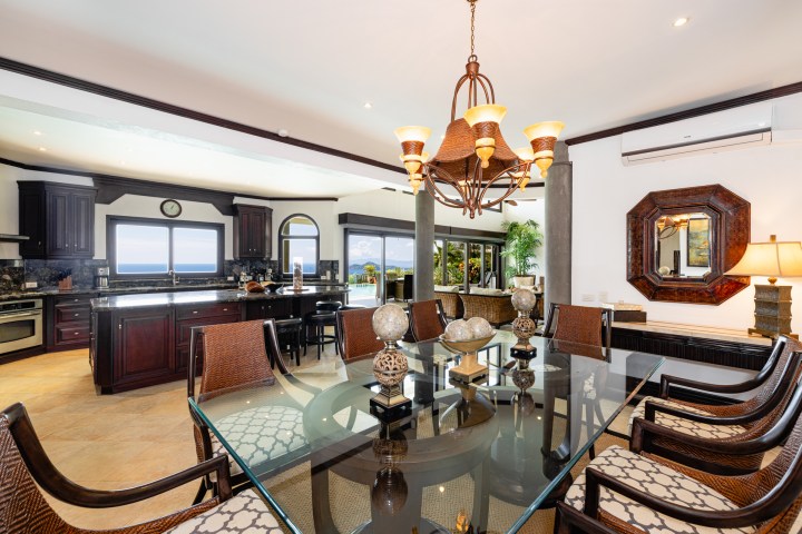 Elegant dining room with glass table, chandelier, and view of modern kitchen with ocean view windows.