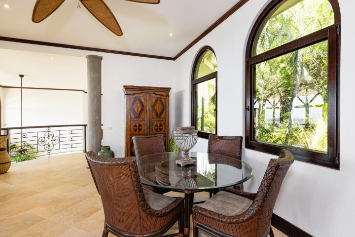Dining area with round glass table, wicker chairs, large windows, and wooden cabinet.
