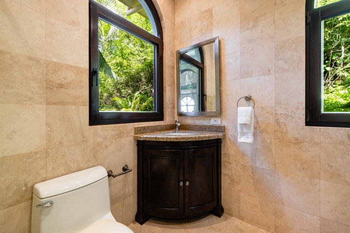 Bathroom with toilet, corner sink, two windows, and beige tiled walls.