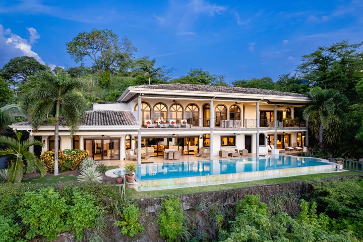 Luxury villa with arched windows, expansive terrace, and infinity pool surrounded by lush greenery.