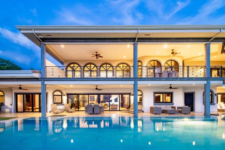 Modern villa with arched windows, balconies, and a pool at sunset.