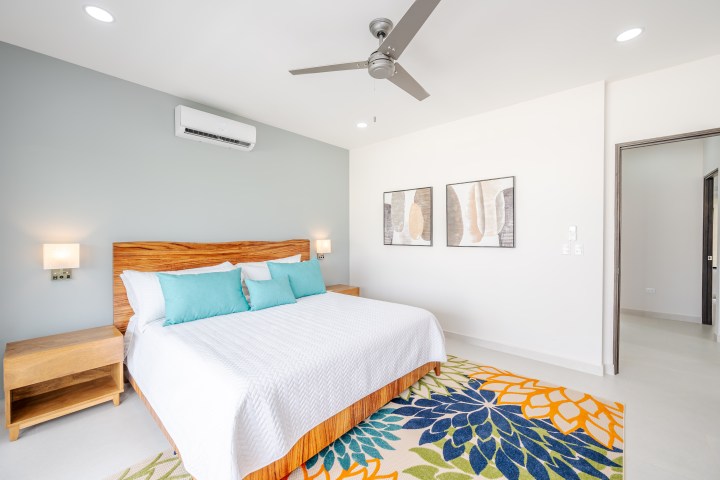 Modern bedroom with wooden bed, teal pillows, and colorful rug under a ceiling fan.
