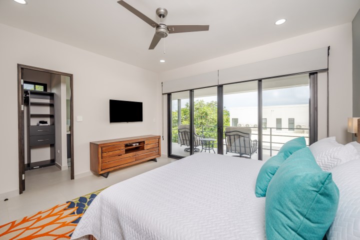 Modern bedroom with balcony, bed with turquoise pillows, wooden TV stand, ceiling fan, and sliding glass doors.