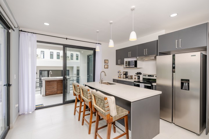 Modern kitchen with gray cabinets, island with chairs, stainless steel appliances, and sliding glass doors.