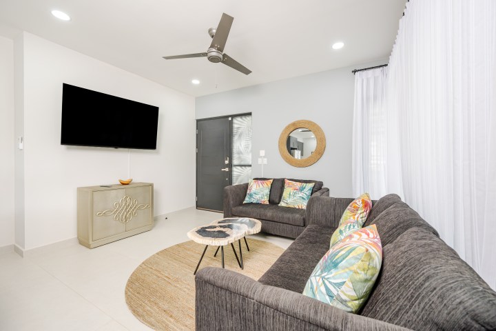 Modern living room with gray sofas, colorful cushions, TV, round mirror, and ceiling fan.