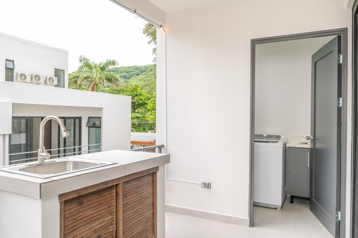 Modern balcony with sink, open door to laundry room, view of trees and building.