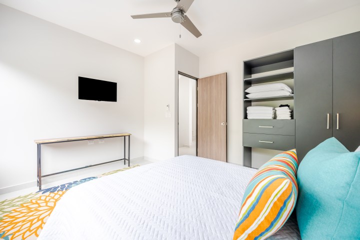 Modern bedroom with bed, colorful pillows, TV, ceiling fan, and open closet with shelves.