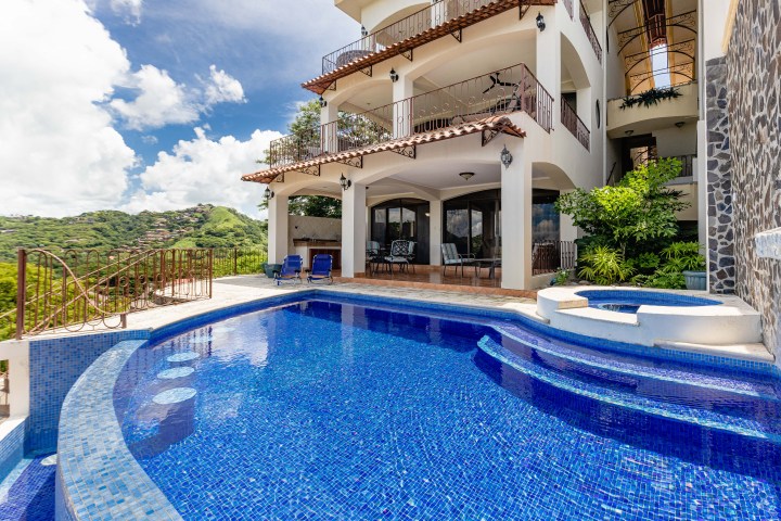 Luxury villa with three levels, balconies, and a blue tiled pool overlooking a lush green landscape.
