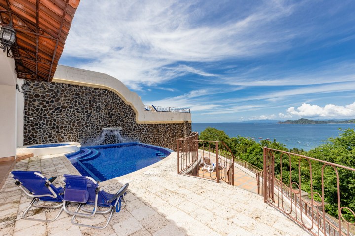 Infinity pool with stone wall waterfall and ocean view, two lounge chairs on patio.