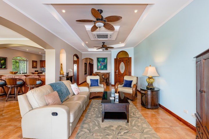 Cozy living room with beige sofas, a ceiling fan, and an open kitchen in the background.