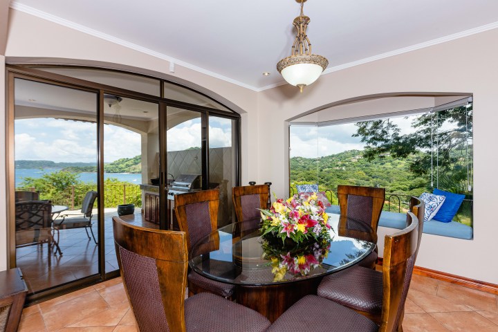 Dining room with glass table, floral centerpiece, large windows showing a scenic view of hills and water.