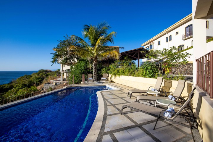 Poolside view with lounge chairs, palm trees, and a glimpse of the ocean and villa.