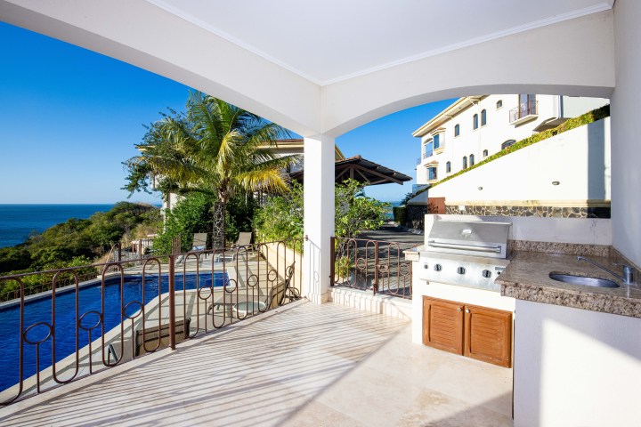 Outdoor patio with grill, sink, ocean view, pool, palm tree, and villa in the background.