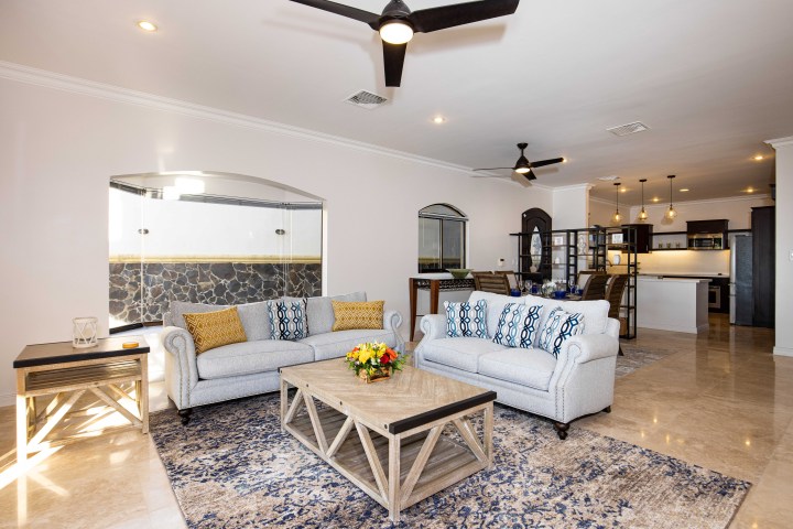 Modern living room with sofas, wooden table, ceiling fans, and open kitchen.