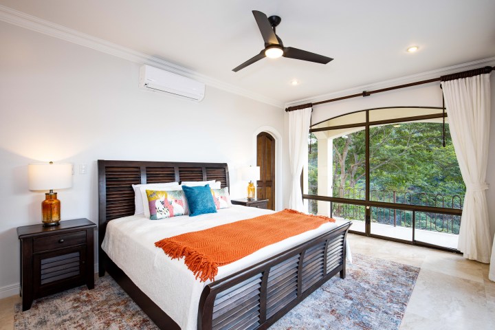 Bright bedroom with a large window, bed with orange throw, pillows, and nightstands with lamps.