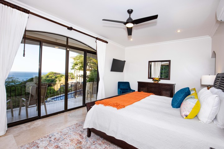 Bright bedroom with ocean view, white curtains, ceiling fan, and colorful pillows on bed.